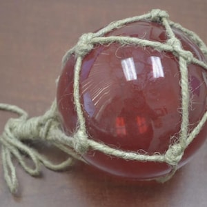 Op de afbeelding: Een rode glazen drijver met een net van natuurlijke touw. De drijver is rond en heeft een touw eraan bevestigd.