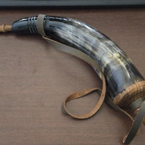 May include: A curved drinking horn featuring a polished black and white marbled surface, a wooden stopper at the narrow end, and a wooden base cap. The horn is fitted with a grey leather strap for carrying and a small wooden plug at the tip.