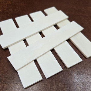 May include: A white, rectangular coaster with a woven design. The coaster is made of several flat, white pieces of wood arranged in a lattice pattern. The coaster is on a brown surface.