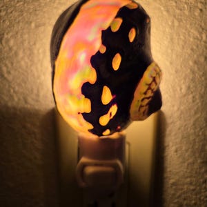 May include: A decorative night light shaped like a skull, featuring a black base with vibrant orange, yellow, and pink accents. The night light is plugged into a standard wall outlet, casting a warm glow.