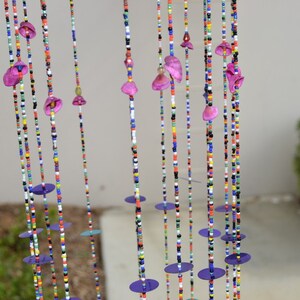 May include: A colorful beaded curtain with purple flower-shaped beads and round purple sequins. The curtain is hanging in front of a white wall.