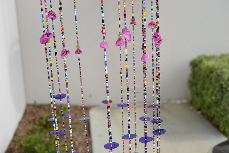 May include: A colorful beaded curtain with purple flower-shaped beads and round purple sequins. The curtain is hanging in front of a white wall.