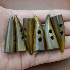 May include: Six wooden toggle buttons in a natural, warm brown hue with a glossy finish. Each button features two or three holes for sewing and a rounded, conical form. The buttons are arranged in a row.