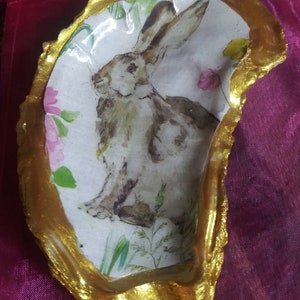 May include: A gold-rimmed oyster shell with a painted image of a rabbit in a field of flowers. The rabbit is brown and white with long ears. The flowers are pink, yellow, and green.