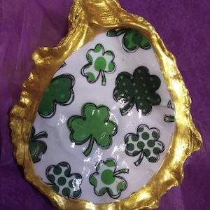 May include: A gold-painted oyster shell with a white interior decorated with a green shamrock pattern. The shamrocks have white outlines and some have white polka dots.
