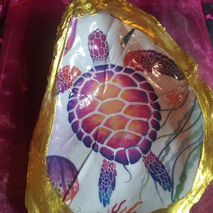 Peut inclure: Un coquillage blanc avec un bord doré, peint avec une tortue de mer colorée et un motif de récif corallien.