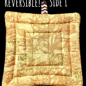 May include: A square quilted potholder in shades of yellow and tan featuring a sunflower print. The text REVERSIBLE! SIDE 1 is printed above the item. A brown and white chevron patterned fabric loop is attached to the top center edge.
