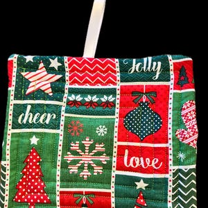 May include: A square quilted potholder featuring a patchwork design of red and green holiday patterns, including Christmas trees, snowflakes, ornaments, and stars. The fabric includes the printed words Jolly, cheer, and love.