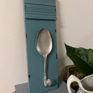 May include: A wall-mounted hook made from a silver-toned metal spoon bent into a hook shape, attached to a rectangular, distressed blue wooden board with a decorative molded top edge and visible black paint wear along the edges.