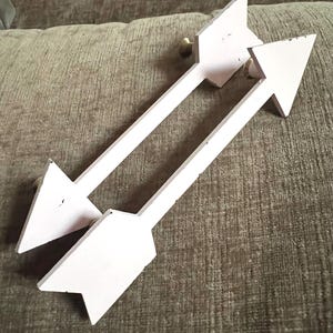 May include: Two wooden arrows painted white with a light pink hue. The arrows have a classic arrowhead shape and are mounted with small, gold-colored knobs. These decorative arrows are likely for wall decor.