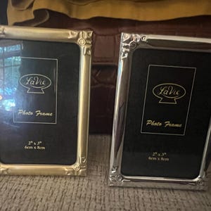 May include: Two rectangular photo frames, one with a gold-tone finish and one with a silver-tone finish. Each frame features decorative corner detailing and a black insert with the text LaVie Photo Frame 2" x 3" 6cm x 8cm printed in gold.