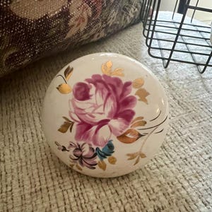 May include: A round, white ceramic knob featuring a hand-painted floral design. The center displays a large, vibrant pink rose surrounded by smaller purple and blue flowers. Delicate gold leaf accents are painted around the floral arrangement.