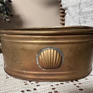 May include: A vintage, oval-shaped brass planter with two handles and a seashell accent. The planter has a weathered, antique appearance, with a tarnished finish. The planter is approximately 12 inches long and 6 inches wide.