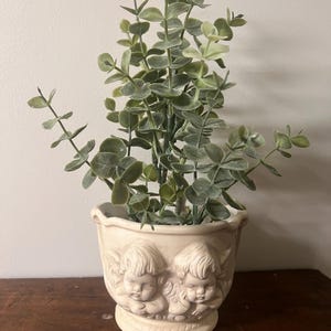 May include: A cream-colored ceramic planter featuring two sculpted cherub faces in relief on the front. The planter contains a faux eucalyptus plant with multiple green stems and rounded leaves extending upward from the center of the vessel.