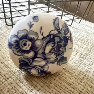 May include: A spherical ceramic knob featuring a white base with a blue floral pattern. The design includes detailed illustrations of blooming flowers and leaves in varying shades of blue, covering the rounded surface of the decorative object.