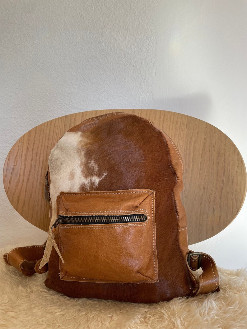 May include: A brown leather backpack with a white and brown cowhide pattern. The backpack has a zippered front pocket and adjustable straps.