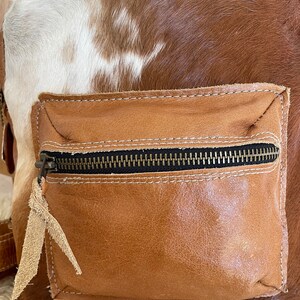 May include: A brown and white cowhide leather backpack with a brown leather front pocket and a zipper closure.