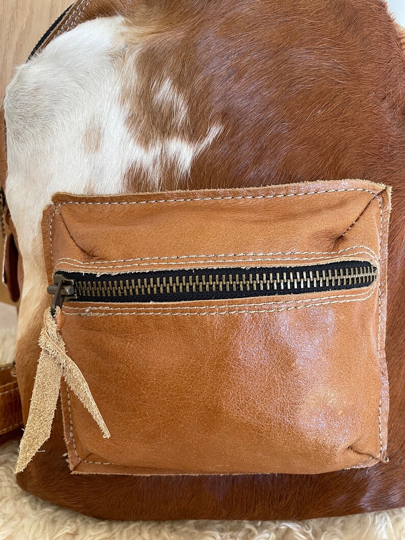 May include: A brown and white cowhide leather backpack with a brown leather front pocket and a zipper closure.