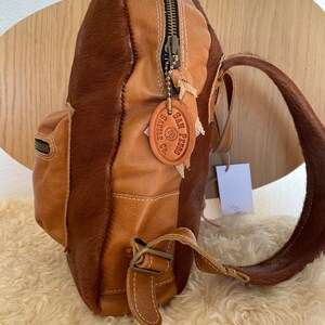 May include: A brown leather backpack with a cowhide patch that says "SAN PEDRO SADDLE CO.". The backpack has a zipper closure and a buckle strap.