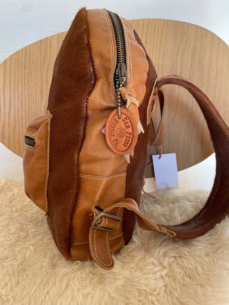 May include: A brown leather backpack with a cowhide patch that says "SAN PEDRO SADDLE CO.". The backpack has a zipper closure and a buckle strap.