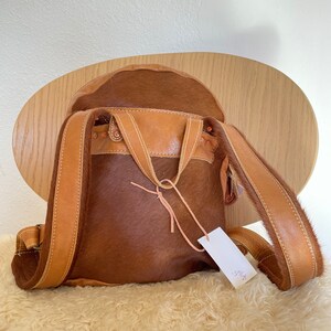 May include: A brown leather backpack with a cowhide exterior. The backpack has a flap closure with a buckle and a small tag that says "$195".