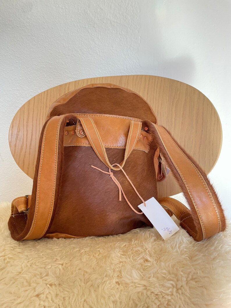 May include: A brown leather backpack with a cowhide exterior. The backpack has a flap closure with a buckle and a small tag that says "$195".