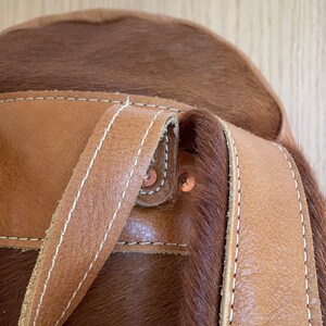 May include: Close-up of a brown leather backpack with a cowhide patch and white stitching. The backpack has two straps and copper rivets.