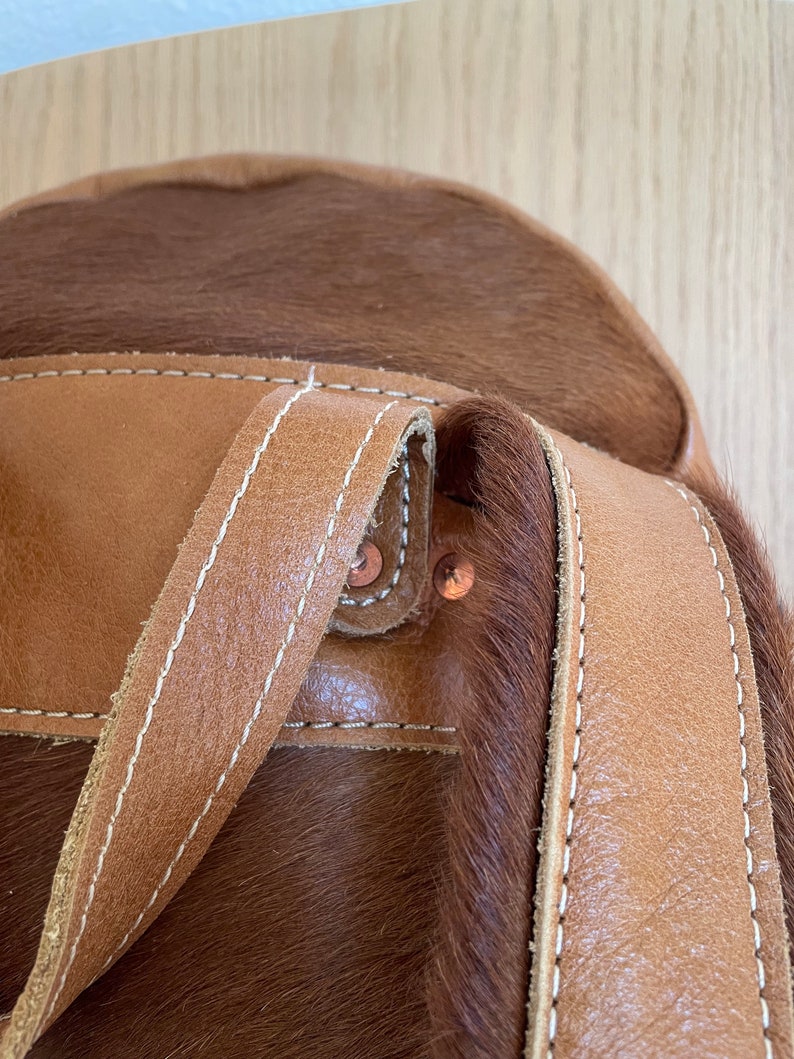 May include: Close-up of a brown leather backpack with a cowhide patch and white stitching. The backpack has two straps and copper rivets.