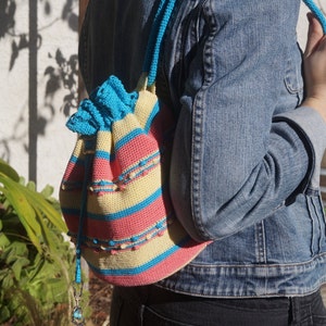 Puede incluir: Una bolsa de cordón de crochet con rayas amarillas, rosas y azules. La bolsa se lleva como una mochila.