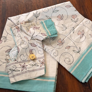 May include: An off-white saree and matching blouse featuring black embroidered vine patterns with small peach-colored flowers and a wide teal border. A small yellow circular measuring tape is placed on the blouse fabric.