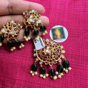 May include: A pair of gold earrings with pink and white stones, green beads, and pearl accents. The earrings are in the shape of a peacock with its tail feathers spread out. The earrings are on a pink surface with a small white sticker that says "Pendant Set 45".