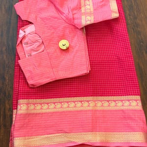May include: A bright red saree featuring a fine white grid pattern and a wide border with gold paisley motifs. The matching light pink blouse has short sleeves, a white grid pattern, and a gold trim. A small gold circular button rests on the blouse.