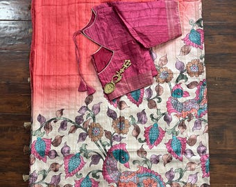 May include: A vibrant, floral-patterned saree with a matching blouse. The saree features a gradient of pink and peach hues, with a detailed border and tassels. The blouse is a deep magenta color, and the saree has a peacock and floral design.