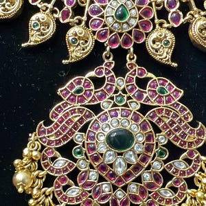 May include: A gold necklace with an intricate design featuring pink and green gemstones. The necklace has a pendant with a floral motif and a pair of peacocks.