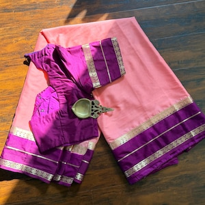 May include: A peach-colored saree featuring a wide purple border with gold metallic trim, paired with a matching purple short-sleeved blouse that has gold metallic bands on the sleeves and a small metallic decorative object placed on top.