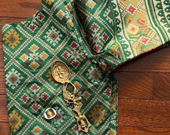 May include: Green patterned fabric with a geometric design, featuring red and yellow accents. A gold-coloured decorative spoon and a small pin are placed on the fabric. The fabric appears to be part of a garment.