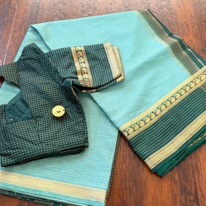 May include: A light turquoise saree with a subtle grid pattern and a dark green border featuring a gold paisley design. A matching dark green blouse with a white grid pattern and gold trim is folded on top of the fabric, featuring a round gold button.