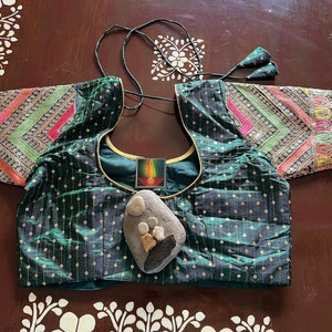 May include: A green, sequined, embroidered blouse with gold trim and colourful patterned sleeves. A small, decorative stone with painted details is placed on the front of the blouse.