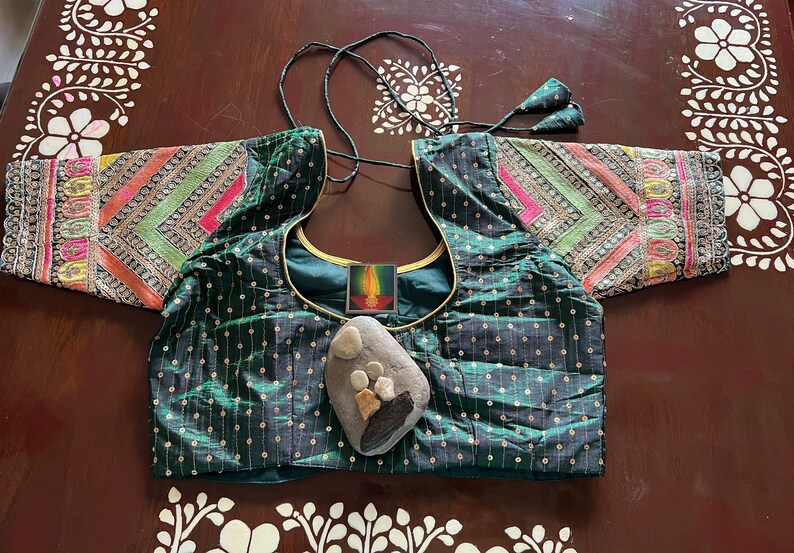 May include: A green, sequined, embroidered blouse with gold trim and colourful patterned sleeves. A small, decorative stone with painted details is placed on the front of the blouse.