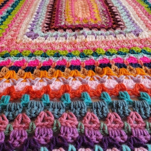 May include: A colourful crocheted blanket featuring a range of bright colours, including pink, orange, green, blue, and purple. The blanket is crafted with a textured stitch that creates a unique and interesting pattern.