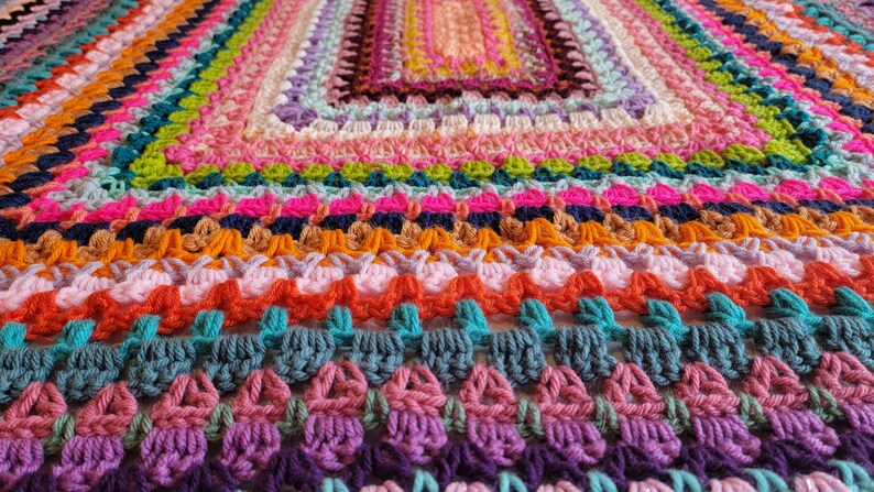 May include: A colourful crocheted blanket featuring a range of bright colours, including pink, orange, green, blue, and purple. The blanket is crafted with a textured stitch that creates a unique and interesting pattern.