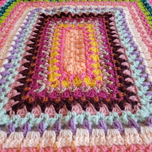 May include: A colourful crocheted blanket with a rectangular pattern in the centre. The blanket is made with a variety of colours, including pink, purple, green, yellow, and orange.