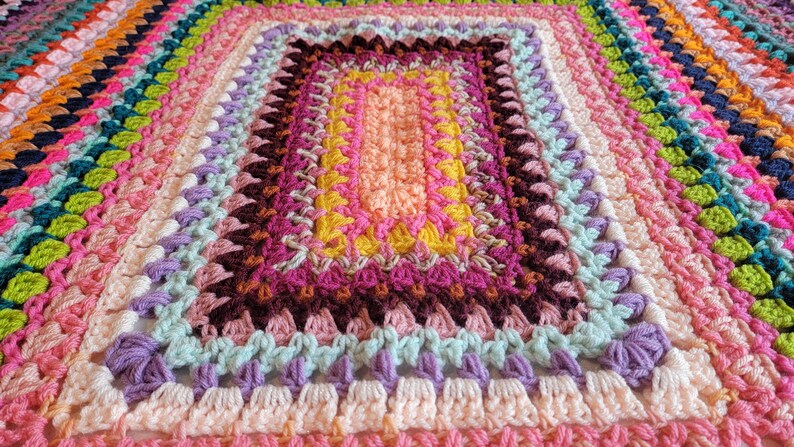 May include: A colourful crocheted blanket with a rectangular pattern in the centre. The blanket is made with a variety of colours, including pink, purple, green, yellow, and orange.