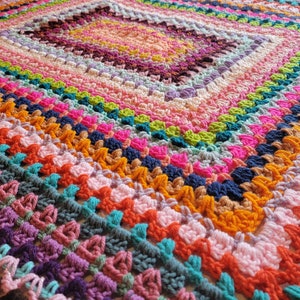 May include: A colourful crocheted blanket with a square pattern in the centre. The blanket is made with a variety of different coloured yarns, including pink, purple, blue, green, orange and yellow.