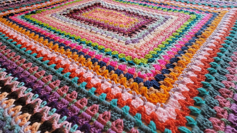May include: A colourful crocheted blanket with a square pattern in the centre. The blanket is made with a variety of different coloured yarns, including pink, purple, blue, green, orange and yellow.