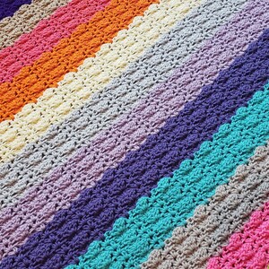 Op de afbeelding: Een kleurrijk gehaakt deken met strepen in turquoise, roze, groen, grijs, paars, crème, oranje en bruin.