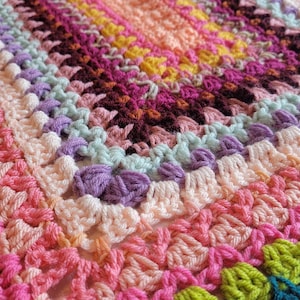 May include: A colourful crocheted blanket featuring a range of bright colours, including pink, green, purple, and blue. The blanket is crafted with a textured stitch and has a scalloped edge.