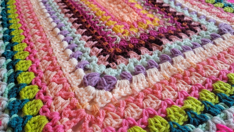 May include: A colourful crocheted blanket featuring a range of bright colours, including pink, green, purple, and blue. The blanket is crafted with a textured stitch and has a scalloped edge.