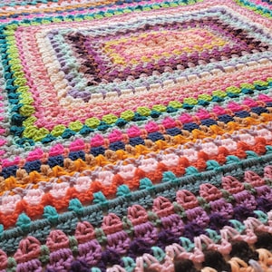 May include: A colourful crocheted blanket with a square pattern in the centre. The blanket is made with a variety of different coloured yarns, including pink, purple, blue, green, yellow, and orange.