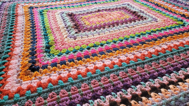 May include: A colourful crocheted blanket with a square pattern in the centre. The blanket is made with a variety of different coloured yarns, including pink, purple, blue, green, yellow, and orange.